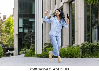 Young joyful beautiful woman dancing and singing in headphones while walking in the city, Hispanic woman with curly hair uses an application on the phone, to listen to music online. - Powered by Shutterstock