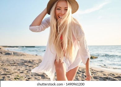 Young joyful blond woman in swimsuit and white shirt wearing hat happily looking in camera with sea on background