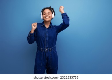 young joyful brunette latin woman dressed in blue denim overalls listens to music in wireless headphones on a blue background