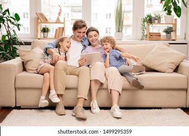 Junge, fröhliche Familie von zwei Kindern und Paaren, die auf dem Sofa sitzen und lustige Videos oder Cartoons auf Touchpad ansehen
