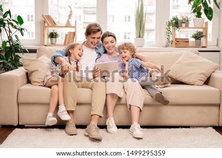 Young joyful casual family of two kids and couple sitting on sofa and watching funny video or cartoons in touchpad