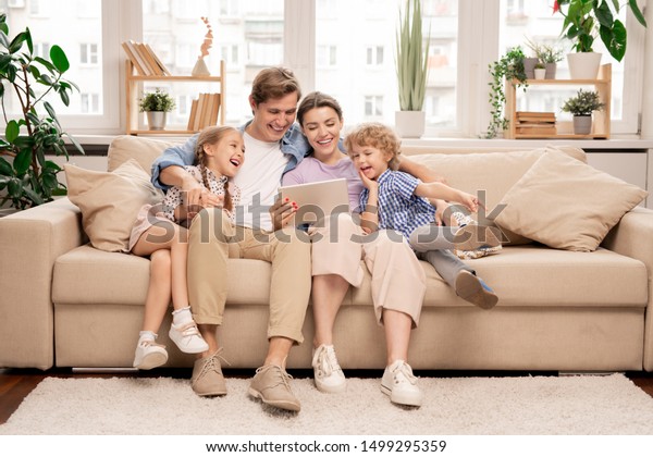 Young joyful\
casual family of two kids and couple sitting on sofa and watching\
funny video or cartoons in\
touchpad