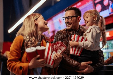 A young joyful Caucasian family is purchasing popcorn, tickets and beverages at the counter in cinema.