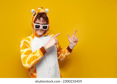 young joyful guy in funny baby giraffe pajamas and glasses points with his hands to the side on yellow background, man in animal cosplay clothes advertises copy space, pajama party concept