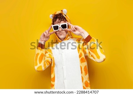 young joyful guy in funny baby giraffe pajamas and glasses is dancing on yellow background, man in animal cosplay clothes shows peace gesture, pajama party concept