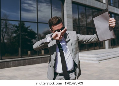 Young joyful happy employee business man in grey suit stand near office glass wall building outdoors in downtown city center hold black folder for paper document cover eye with victory v-sign gesture