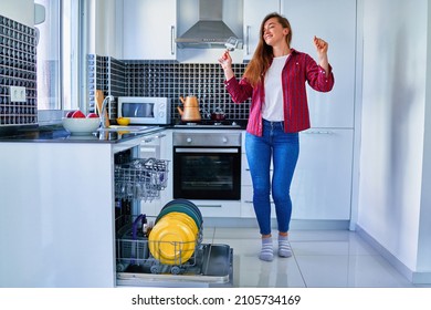 Young joyful happy satisfied smiling cute dancing housewife woman using modern dishwasher for wash dishes at white home kitchen. Enjoy cleaning chores 