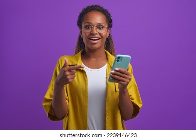 Young joyful inspired African American woman points to phone and sighs with smile after reading good news about career advancement or getting grant at university stands on plain lilac background