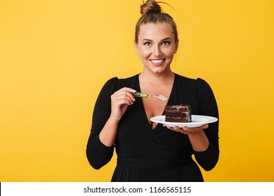 Young joyful woman in black dress happily looking in camera holding plate with half of chocolate cake in hands over yellow background. Plus size model - Powered by Shutterstock