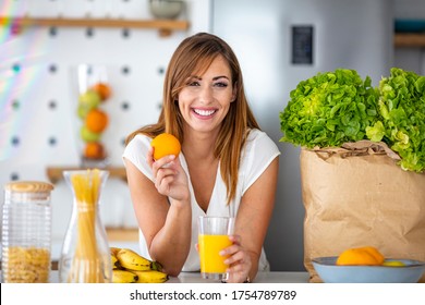 Young joyful woman drinking orange juice and standing near a kitchen table. Close up of a woman drinking juice in her kitchen. Fit smiling young woman preparing healthy fruit juice - Powered by Shutterstock