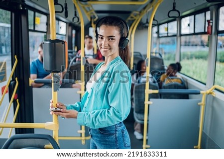 Young joyful woman standing on the bus and using the smartphone and smiling. Public transportation and people concept. Listening music on a headphones and looking into the camera.