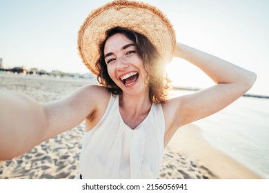Young joyful woman in white shirt wearing hat smiling at camera on the beach - Traveler girl enjoying freedom taking selfie on a sunny day - Wellbeing, healthy lifestyle and happy people concept