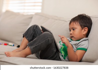 A Young Kid, 3 Years Old Who Is Using His Little Finger On The Touchscreen Of His Big Tablet, While He Watches  The Internet On A Comfy Couch.