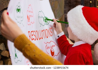 Young Kid Child Boy In Red Santa Hat Making His Resolution Goals For 2019 New Year For His Family: Mother Woman And Father Male Man During Winter Holidays. Merry Christmas And Happy New Year Concept.