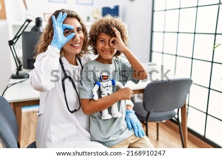 Young kid at pediatrician clinic holding teddy bear smiling happy doing ok sign with hand on eye looking through fingers 