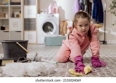 A young kid using a washcloth and liquid at home. Disgruntled girl mopping the apartment floor, doing housework, playing, front view in the laundry room - concept of child and adolescent development.