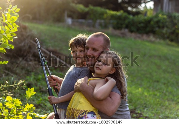 Young kids help out their father in the\
garden,cutting leaves together.Adorable siblings helping their dad\
gardening.A young single father teaching son and daughter how to\
prune a bush in the\
backyard