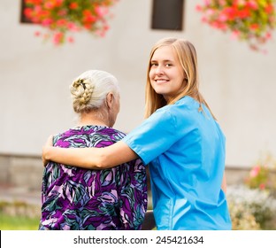 Young Kind Doctor Helping Old Lady On A Walk In The Garden.