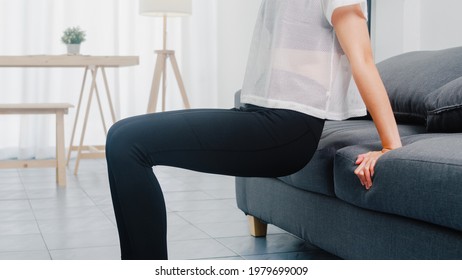 Young Korean Lady In Sportswear Exercises Doing Working Out Doing Tricep Dips Leaning On Couch In Living Room At Home. Social Distance, Isolation During The Virus. Exercises For The Lower Body.