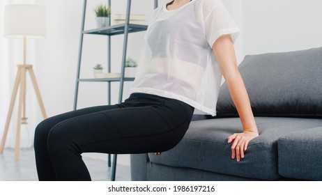 Young Korean Lady In Sportswear Exercises Doing Working Out Doing Tricep Dips Leaning On Couch In Living Room At Home. Social Distance, Isolation During The Virus. Exercises For The Lower Body.