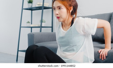 Young Korean Lady In Sportswear Exercises Doing Working Out Doing Tricep Dips Leaning On Couch In Living Room At Home. Social Distance, Isolation During The Virus. Exercises For The Lower Body.