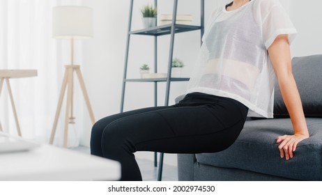 Young Korean Lady In Sportswear Exercises Doing Working Out Doing Tricep Dips Leaning On Couch In Living Room At Home. Social Distance, Isolation During The Virus. Exercises For The Lower Body.