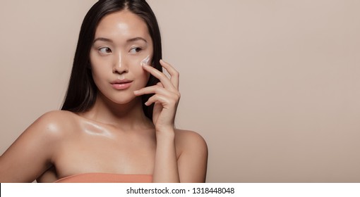 Young Korean woman applying moisturizer to her face over beige background. Young woman with cosmetic cream on a cheek and looking away.