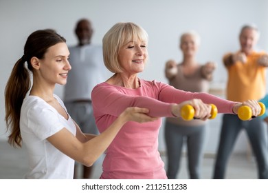 Young lady coach helping senior woman while exercising with dumbbells, sporty multiracial group of elderly people having fitness class at nursing home, training with instructor