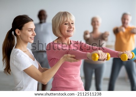 Young lady coach helping senior woman while exercising with dumbbells, sporty multiracial group of elderly people having fitness class at nursing home, training with instructor