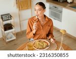 Young lady eating delicious homemade pasta, enjoying tasty lunch with closed eyes while sitting at table in light cozy kitchen interior, free space, above view