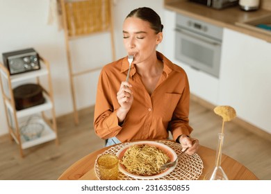 Young lady eating delicious homemade pasta, enjoying tasty lunch with closed eyes while sitting at table in light cozy kitchen interior, free space, above view - Shutterstock ID 2316350855