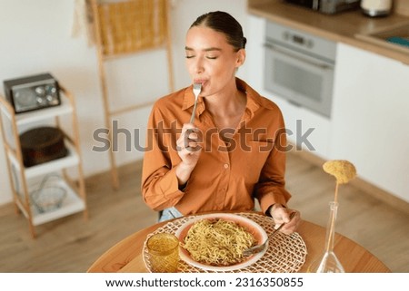 Young lady eating delicious homemade pasta, enjoying tasty lunch with closed eyes while sitting at table in light cozy kitchen interior, free space, above view