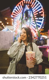 A young lady in a good mood with a popcorn cup in the night at luna park in Santa Monica in front of unfocused illuminations of a Ferris wheel. Santa Monica. California.