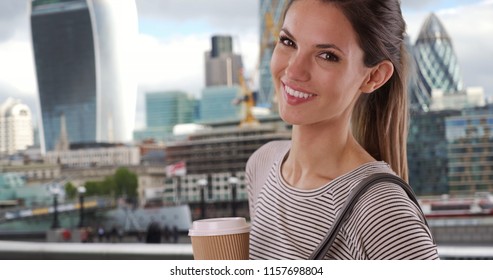 Young lady in her 20s with coffee cup smiling with view of cityscape behind her