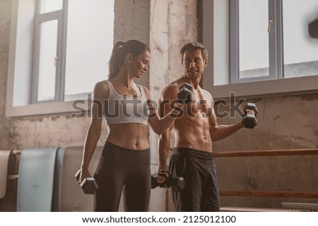 Young lady holding dumbbell and performing exercise for arms with strong athletic man in the gym