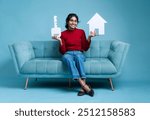 Young lady holding a house model and key, real estate and home loan concept image, Dream home photo, Indian girl sitting on sofa isolated on light blue background