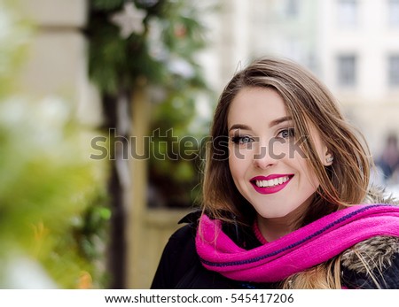 Young lady with long blonde hair and perfect makeup looking at the camera and smiling, outdoor shooting in the city. Winter look in stylish clothes. Light colors photo, close up portrait with green 