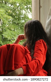 Young Lady With Long Dark Hair, Wearing Red Bathrobe, Sitting And Looking Out The Window. Green Trees Outside. Stay Home During Quarantine