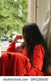 Young Lady With Long Dark Hair, Wearing Red Bathrobe, Sitting And Looking Out The Window. Green Trees Outside. Stay Home During Quarantine