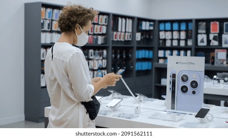 Young Lady In Protective Mask Testing New IPhone In An Electronic Store, Shopping During A Covid-19 Pandemic Outbreak. Denpasar - Indonesia - 2021