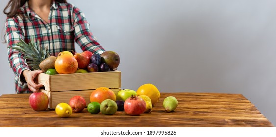 Young Lady Putting Wooden Box With Fruit On The Table. Woman In Flannel Shirt Touching Wood Crate Full Of Fresh Sweet Food On The Brown Desk With Others Plants Around. Person With Tasty Meal.