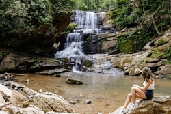 The Young Lady Is Sitting On Large Rocks Next To A High Waterfall And A Mountain River. Beautiful American Landscape With High Waterfall, Mountain River, Forest And People On A Summer Day.