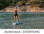 A young lady smiles as she paddles on a stand up paddleboard on calm water of lake or sea in warm evening sunshine; slim woman in swimsuit with shorts is enjoying active water sport on holiday on SUP 