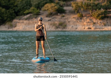 A young lady smiles as she paddles on a stand up paddleboard on calm water of lake or sea in warm evening sunshine; slim woman in swimsuit with shorts is enjoying active water sport on holiday on SUP  - Powered by Shutterstock