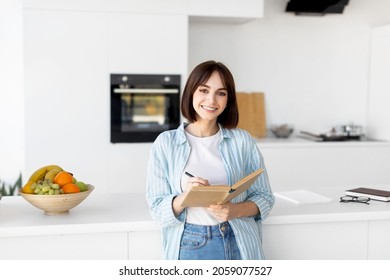 Young lady smiling and taking notes, holding notepad and pen, standing in kitchen interior, copy space. Cheerful millennial woman planning her day, writing tasks - Powered by Shutterstock