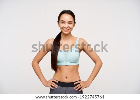 Young lady, sporty asian woman with dark long hair gathered in a ponytail. Wearing sportswear and holding hands on a waist, smiling. Watching at the camera isolated over white background