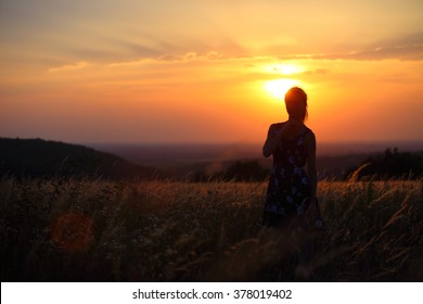 Young Lady Standing Alone In A Field During Sunset. Silhouette Of A Thinking Woman.