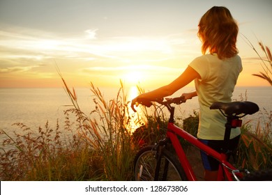 Young Lady Standing With Bicycle On A Sea Coast And Enjoying Sunset