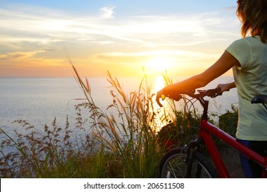 Young lady standing with bicycle on a sea coast and enjoying sunset