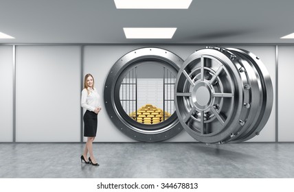 Young Lady Standing In Front Of A Big Unlocked Round Metal Safe In A Bank Depository With A Pyramid Of Gold Bars And Lock-boxes Inside,  A Concept Of Security And Client Service